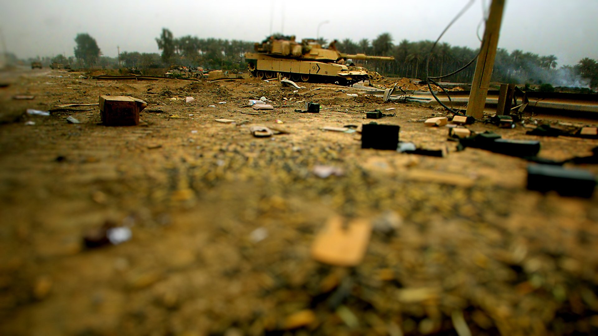 HD PC desktop wallpaper: military tank on a debris-strewn, dusty battlefield with a shallow depth of field and blurred tree line in the background.
