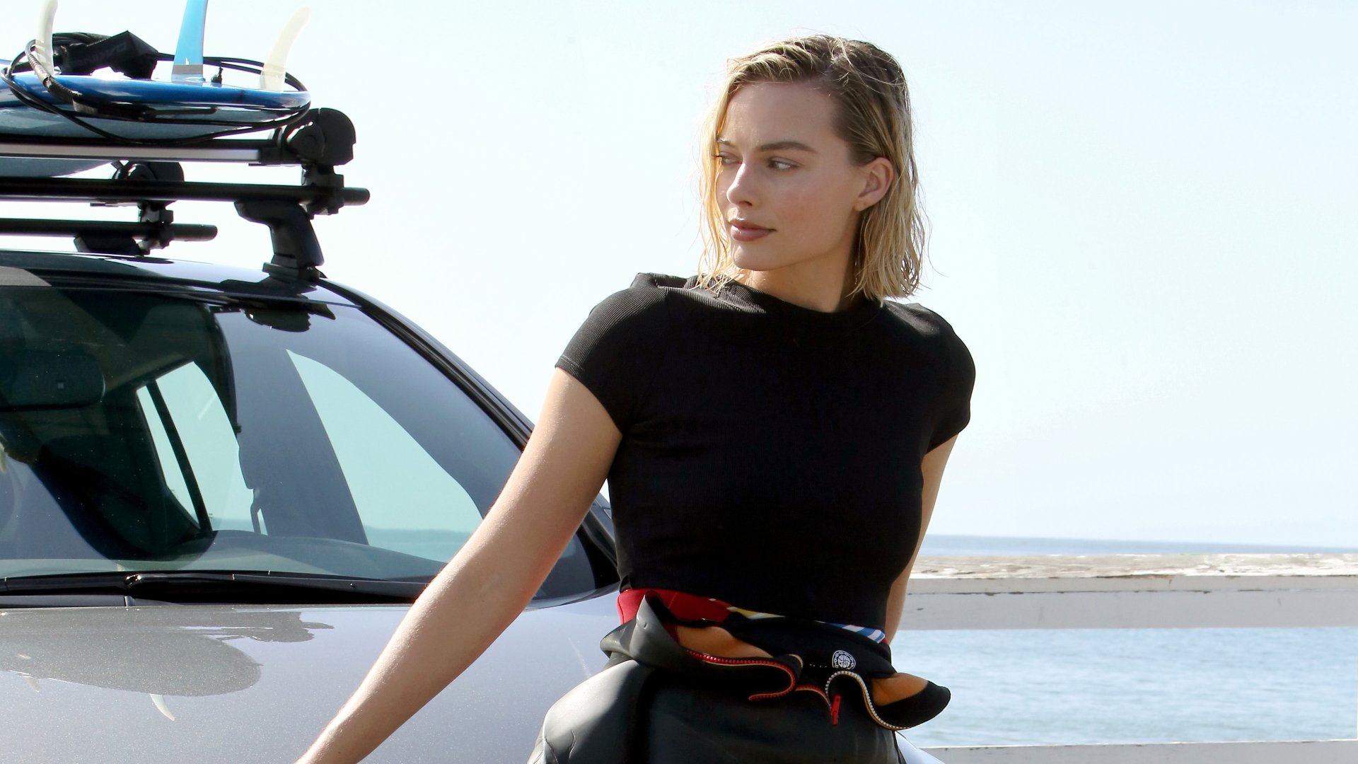 Blonde celebrity Margot Robbie in a casual black outfit leaning against a car by the beach, captured in stunning 4K Ultra HD for PC desktop wallpaper.