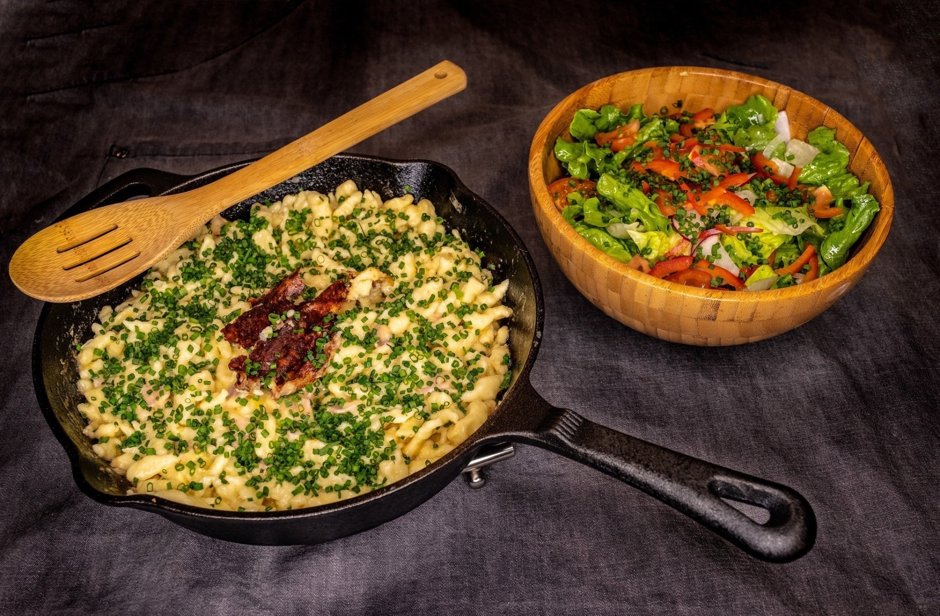 HD PC desktop wallpaper showing Indian food with a cast iron skillet of herb-garnished paneer dish and a wooden bowl of fresh mixed salad on a dark cloth background.