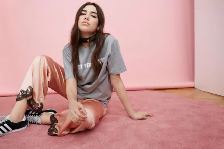 HD desktop wallpaper featuring English brunette singer Dua Lipa sitting on a pink carpet, wearing a casual grey t-shirt and peach-colored pants with black sneakers, set against a pastel pink background.