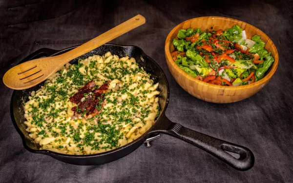 HD PC desktop wallpaper showing Indian food with a cast iron skillet of herb-garnished paneer dish and a wooden bowl of fresh mixed salad on a dark cloth background.