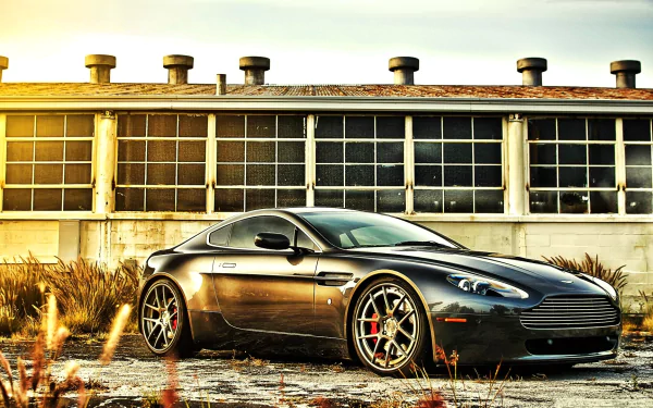 A sleek black Aston Martin DB9 grand tourer parked in front of an industrial building, showcased as a high-definition PC desktop wallpaper and background.