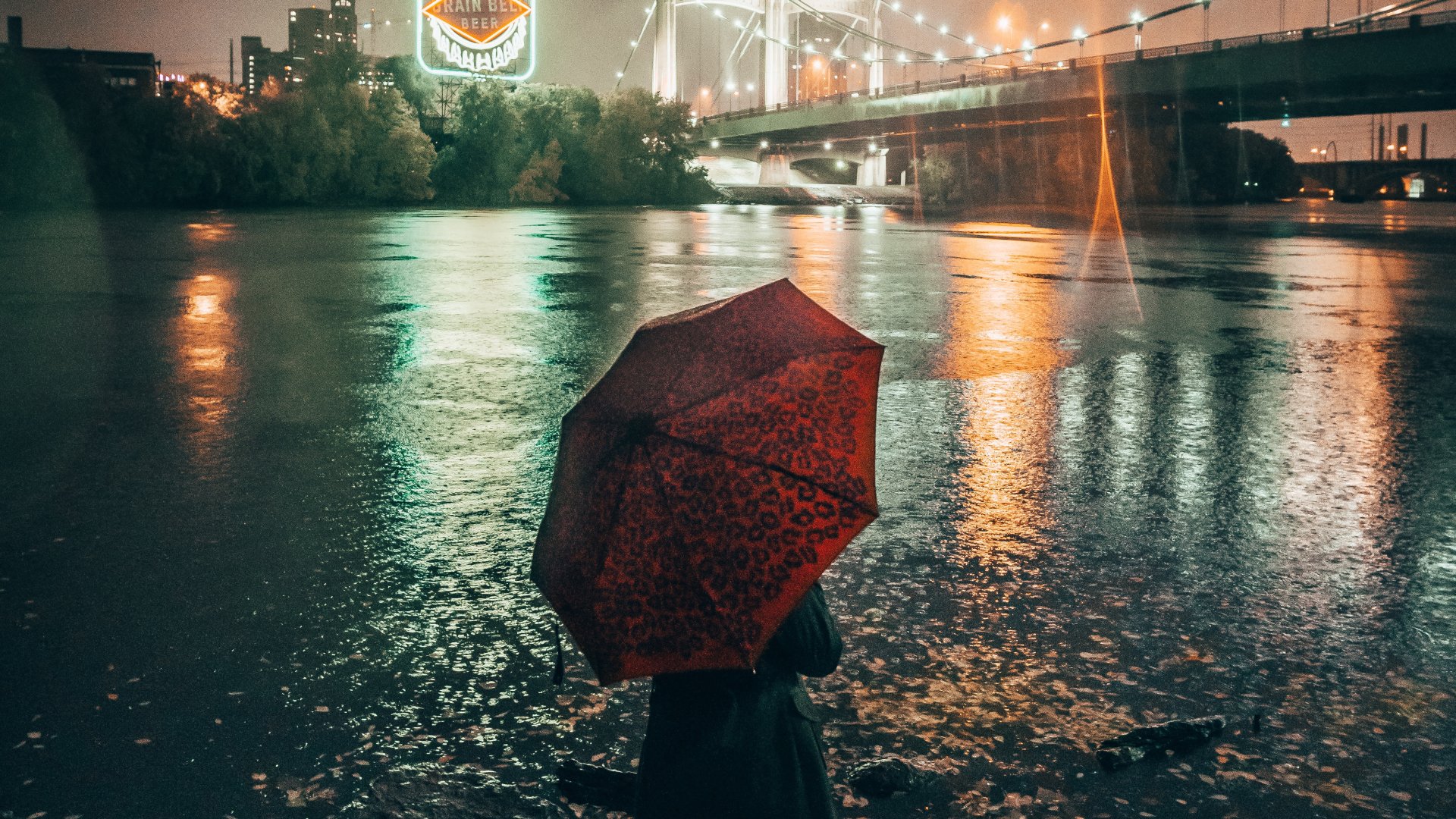A woman holding a red umbrella stands by a river at night, with a lit bridge and city lights reflected in the water, captured in 4K Ultra HD.