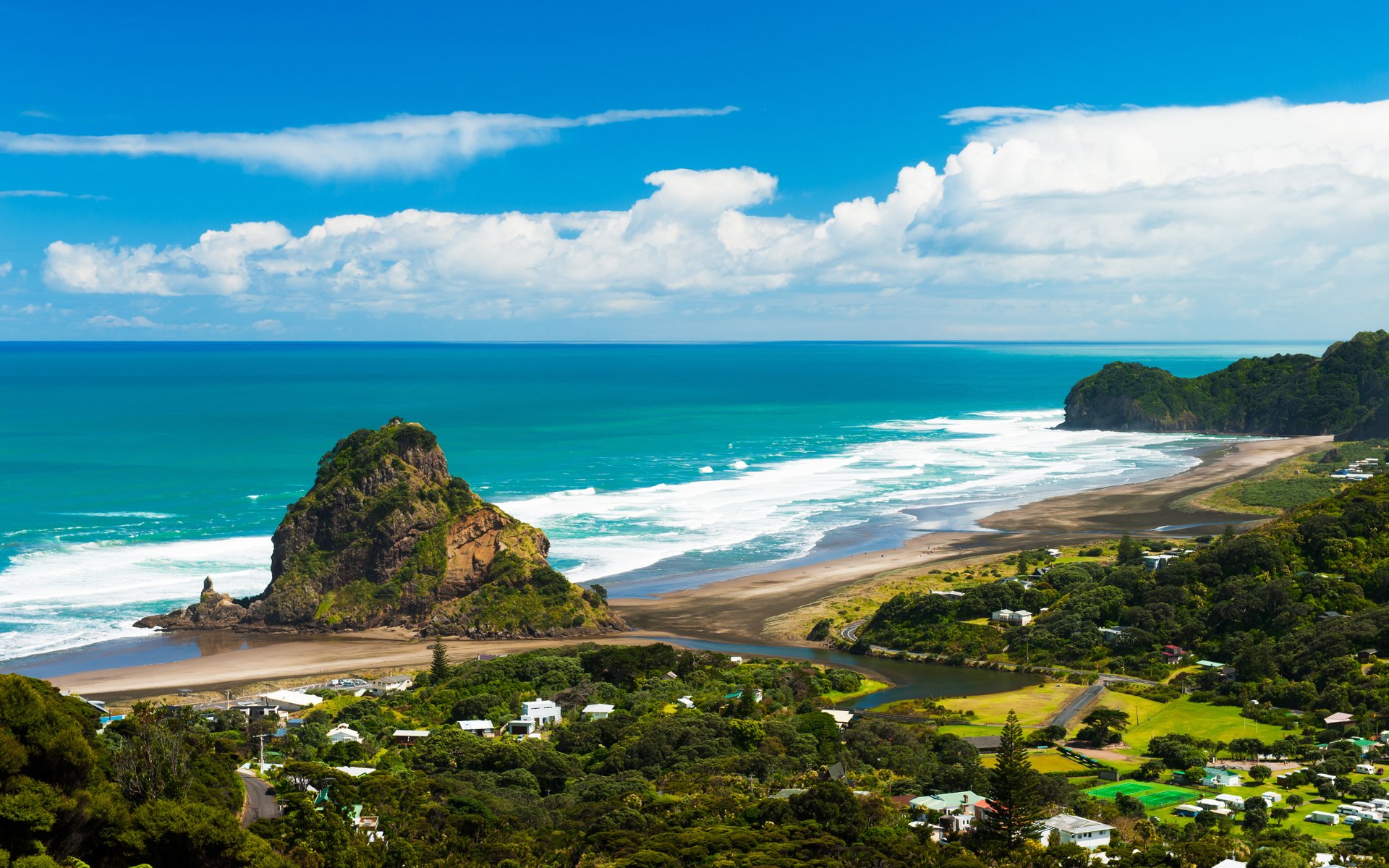 4K Ultra HD view of a New Zealand coastline featuring a coastal village, lush greenery, and ocean waves under a bright blue sky with scattered clouds.