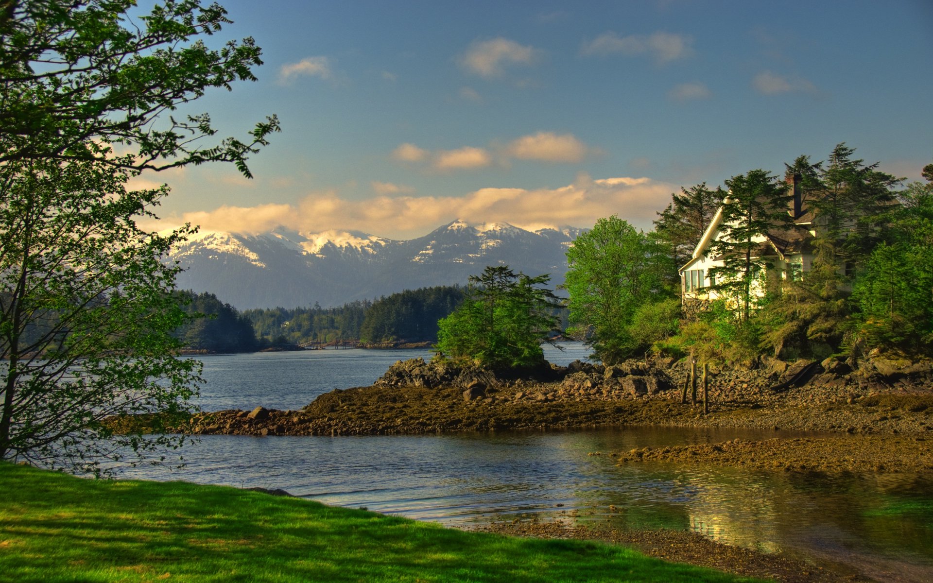 A serene Alaska landscape featuring a lake surrounded by lush forests and snow-capped mountains, with a charming home nestled among the trees, shrouded in gentle clouds.