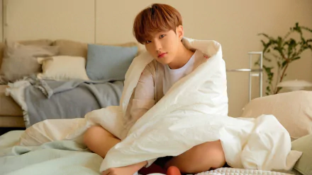 4K PC desktop wallpaper of a K-pop singer wrapped in a white duvet on a bed, soft morning light and cozy music-themed mood.