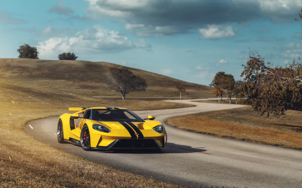 A yellow Ford GT with black racing stripes drives along a winding road under a partly cloudy sky, captured in stunning 8K Ultra HD resolution for a PC desktop background.