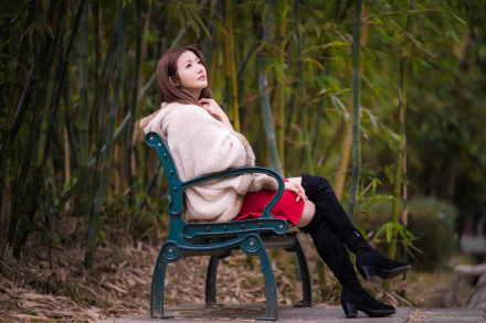 Brunette woman model in a red dress with black knee-high boots seated on a park bench among bamboo — 5K Ultra HD PC desktop wallpaper/background.