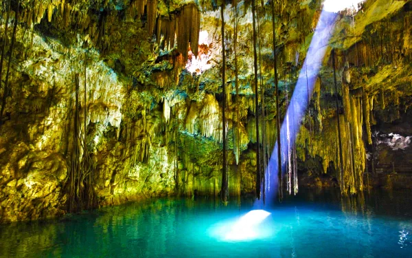 HD nature desktop wallpaper featuring a sunbeam piercing through a cave opening, illuminating turquoise water surrounded by moss-covered rock formations.