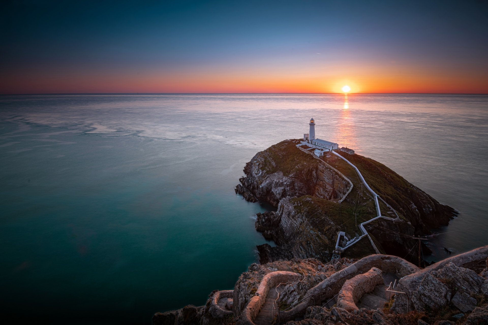 Sunset over the ocean horizon in Wales, featuring a man-made lighthouse on a rocky cliff, captured in a stunning HD desktop wallpaper.