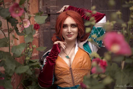 HD PC wallpaper featuring a redhead model cosplaying Triss Merigold from The Witcher 3: Wild Hunt, surrounded by flowers and medieval stonework.