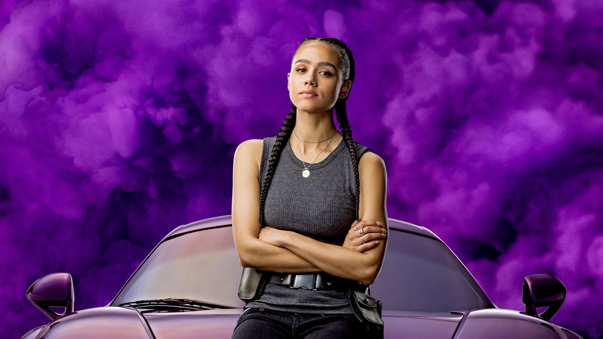 Nathalie Emmanuel as Ramsey from Fast & Furious 9 stands confidently with arms crossed in front of a car against a vibrant purple smoke background.