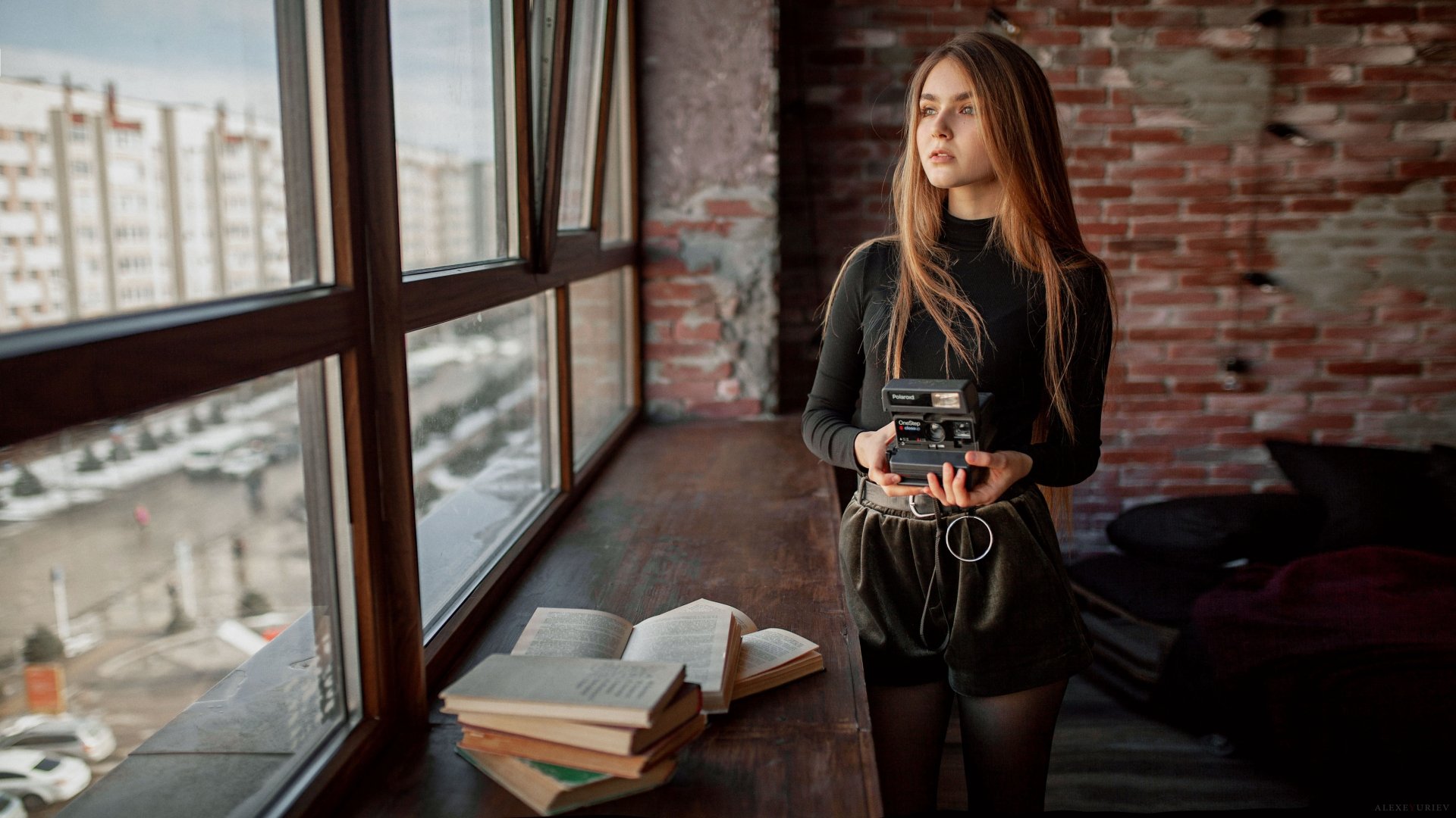 Redhead woman model with long hair holding a camera, standing beside a window in an industrial-style room with books, captured in 4K Ultra HD.