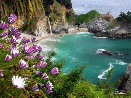 HD PC desktop wallpaper showcasing a serene beach with turquoise waters, rocky cliffs, a small waterfall, and vibrant purple flowers in the foreground.