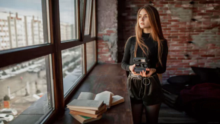 Redhead woman model with long hair holding a camera, standing beside a window in an industrial-style room with books, captured in 4K Ultra HD.