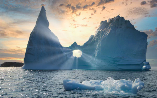4K Ultra HD desktop wallpaper of a sunbeam streaming through a natural hole in a towering iceberg against a colorful sky over the ocean.