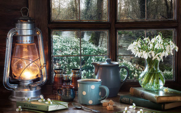 A cozy still life scene with a glowing kerosene lamp, a kettle, a polka-dot mug, books, and a vase of snowdrop flowers by a window showing a snowy garden outside.