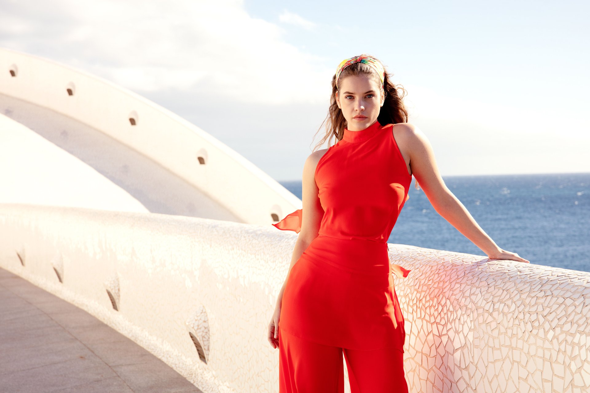 Brunette model in a red sleeveless jumpsuit leaning on a white curved seaside balustrade, sunlit 5K Ultra HD PC desktop background.