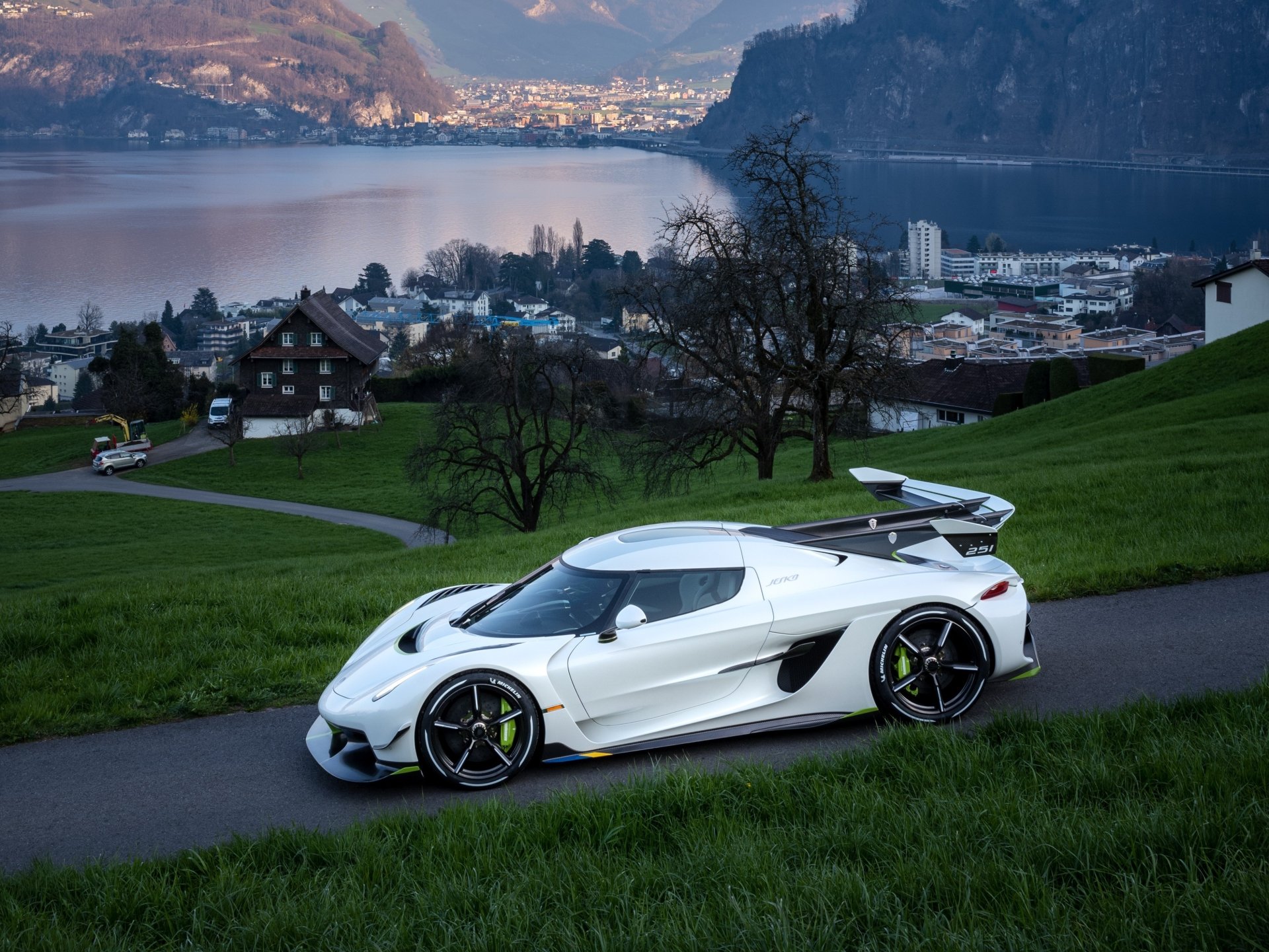 A white Koenigsegg Jesko supercar parked on a winding road with a scenic lakeside town and hills in the background, captured in a high-definition desktop wallpaper.