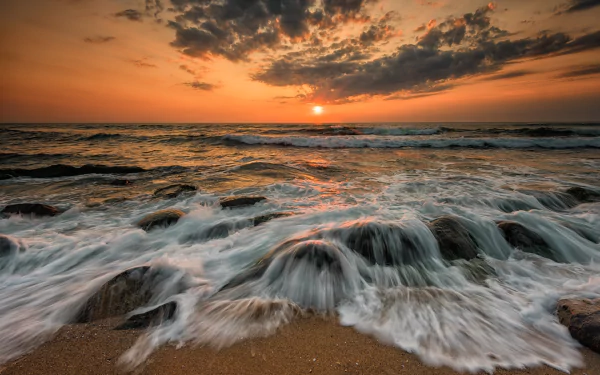 HD desktop wallpaper of an ocean sunset. Waves gently crash over rocks with a vibrant orange sun setting on the horizon, casting warm light over the serene scene. Nature's beauty captured effortlessly.