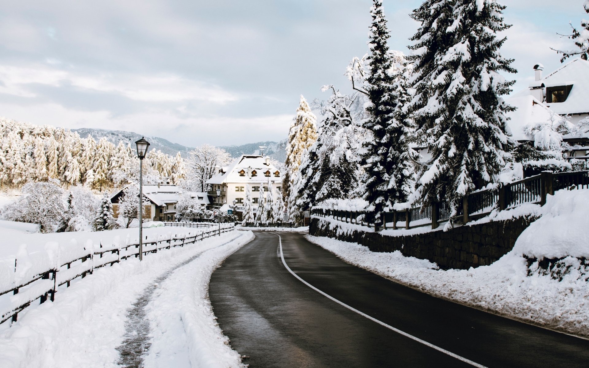 Snow-covered road winding through a winter town in Tyrol, Italy, surrounded by snowy trees and mountains under a cloudy sky.