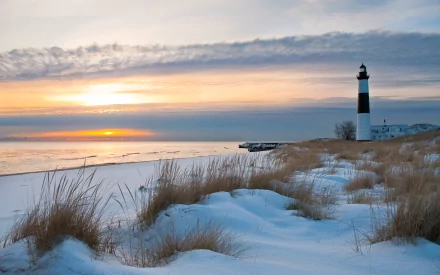 HD desktop wallpaper featuring a picturesque lighthouse standing against a serene winter landscape with a beautiful sunset, snow-covered ground, and tall grass in the foreground.