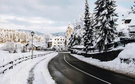 Snow-covered road winding through a winter town in Tyrol, Italy, surrounded by snowy trees and mountains under a cloudy sky.