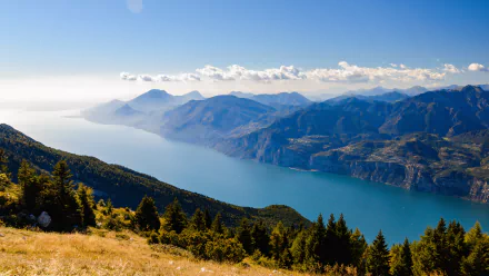 Panoramic 4K Ultra HD view of Lake Garda in Italy, surrounded by the Alps with mountain ridges, lush forests, and clear blue sky in this nature desktop wallpaper.