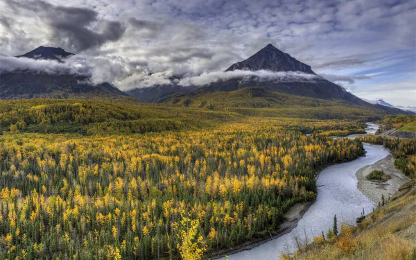 HD desktop wallpaper of a vibrant Alaskan landscape featuring a winding river through a dense forest with autumn colors, set against towering mountains under a cloudy sky.