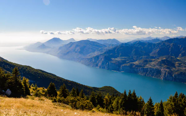 Panoramic 4K Ultra HD view of Lake Garda in Italy, surrounded by the Alps with mountain ridges, lush forests, and clear blue sky in this nature desktop wallpaper.