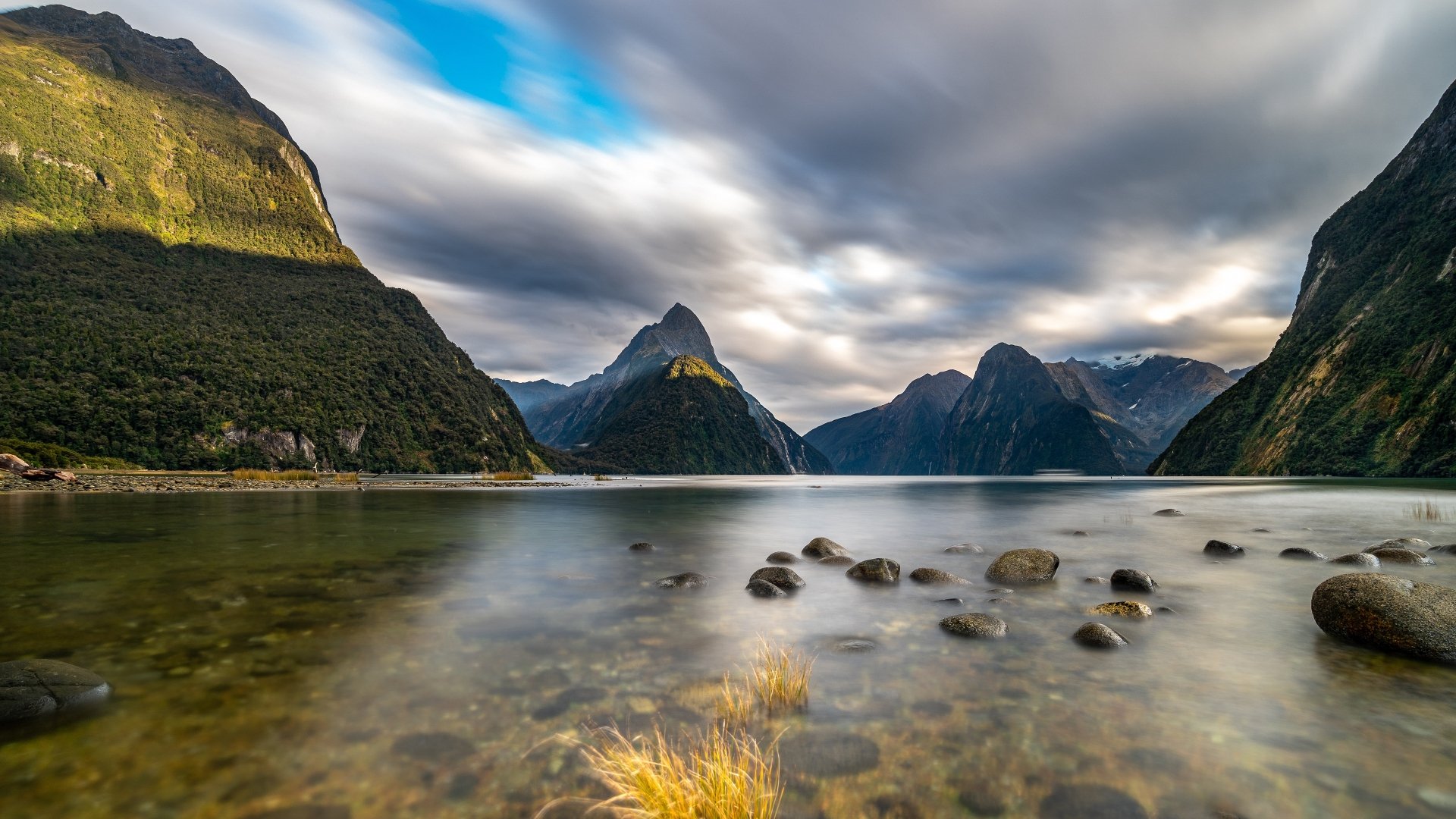 4K Ultra HD desktop wallpaper of a serene lake shore with stones, surrounded by towering mountains under a dynamic cloudy sky in a natural landscape.