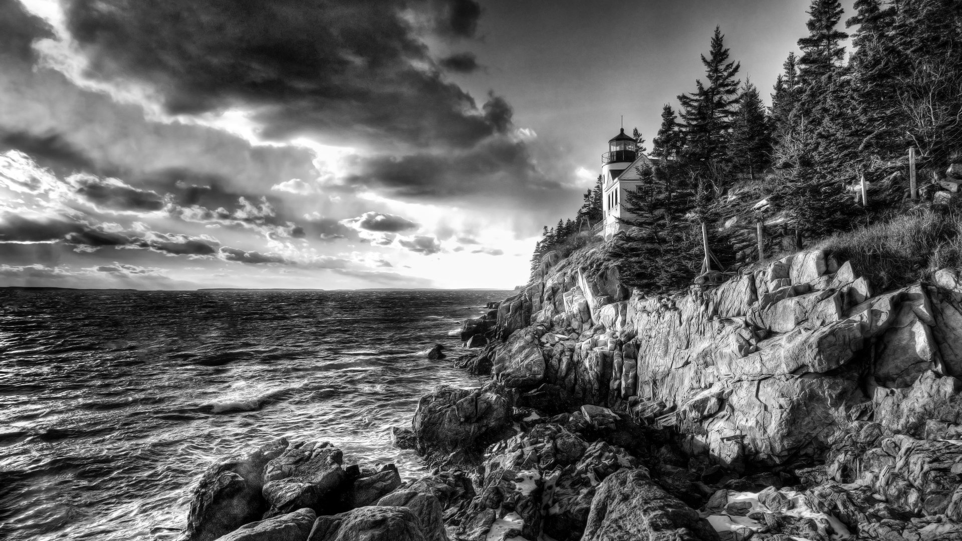 Black and white 4K Ultra HD desktop wallpaper of a lighthouse perched on a rocky coastline with ocean waves and dramatic cloudy sky.