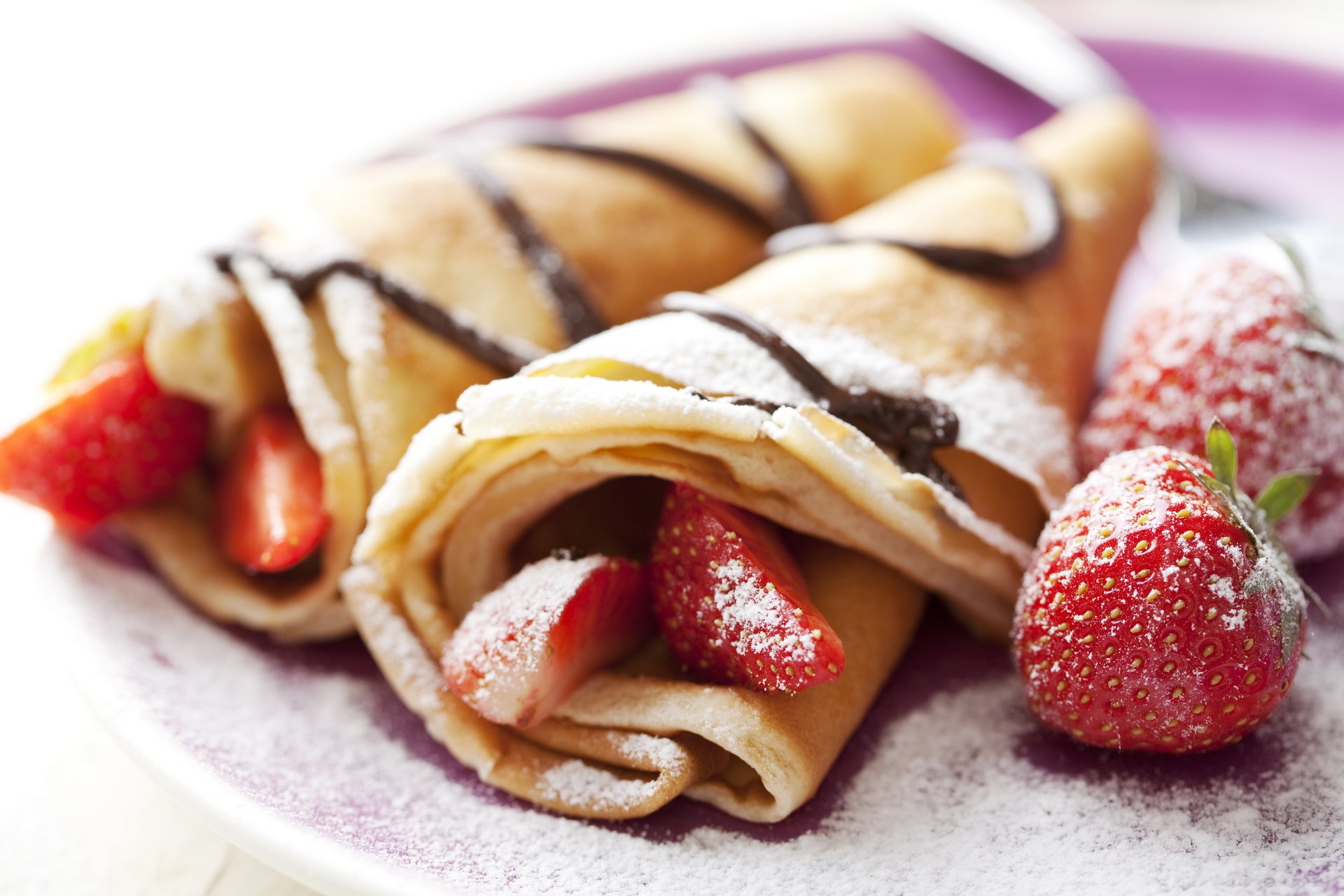 HD desktop wallpaper featuring breakfast crêpes filled with fresh strawberries and drizzled with chocolate, accompanied by whole strawberries on a white plate.