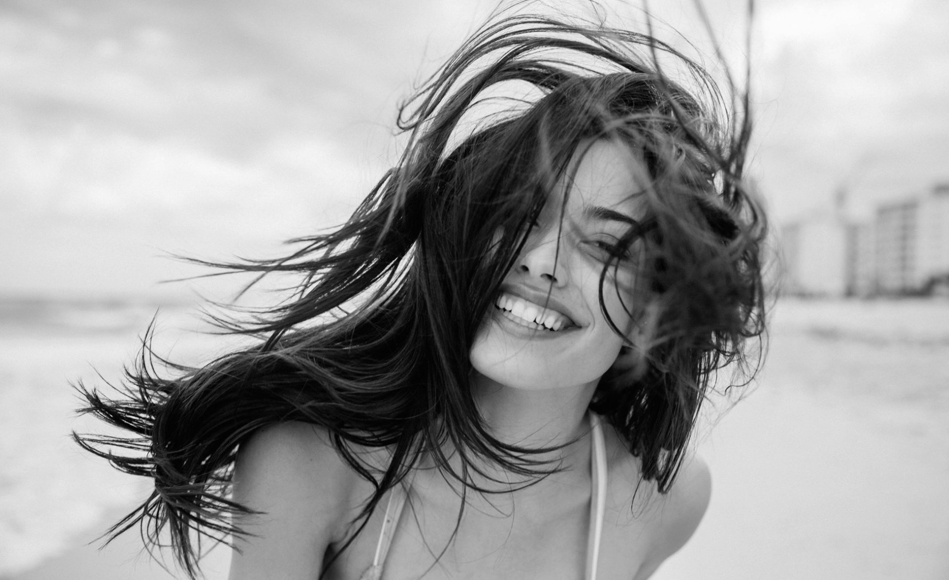 Black-and-white HD PC desktop wallpaper of a smiling woman model on the beach, hair blowing across her face.