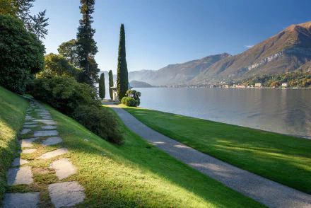 Bellagio, Lake Como, Italy: sunlit lakeside path with cypress trees and mountains reflected in calm water — 2K Quad HD PC desktop wallpaper photography.