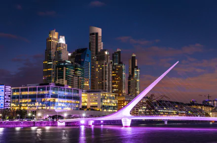 Night view of Buenos Aires cityscape in Argentina, featuring illuminated modern buildings and the glowing Puente de la Mujer bridge over the water.
