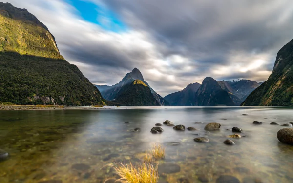 4K Ultra HD desktop wallpaper of a serene lake shore with stones, surrounded by towering mountains under a dynamic cloudy sky in a natural landscape.