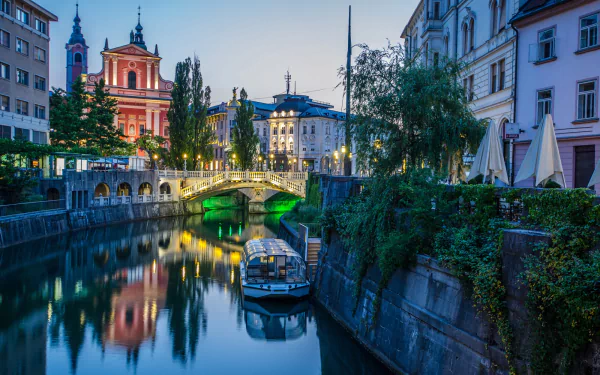 HD PC desktop wallpaper of Ljubljana, Slovenia: evening city river view with boat, arched bridge, church steeples and man-made riverside buildings.