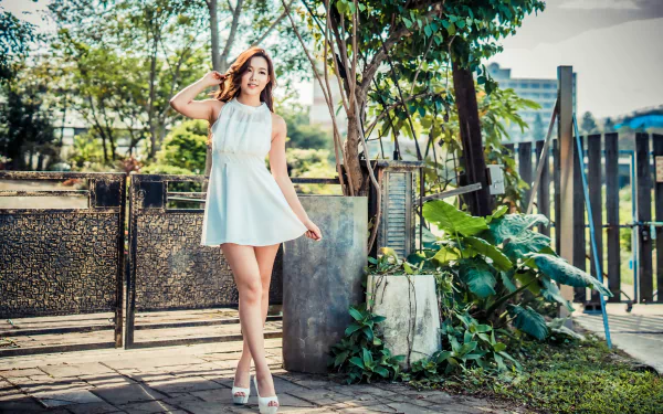 Brunette Asian woman in a white dress poses outdoors by a gate and greenery, captured in 4K Ultra HD for a vibrant PC desktop wallpaper background.