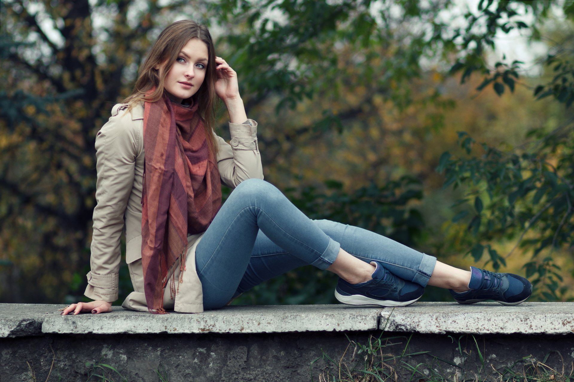 Brunette woman wearing a rust-colored scarf and casual outfit, sitting on a concrete ledge with a forest background, captured in a 4K Ultra HD desktop wallpaper style.