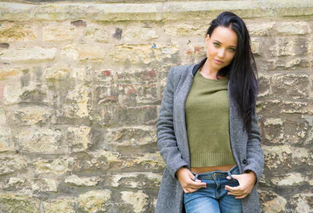 Ukrainian model Angelina Petrova with black hair and green eyes poses confidently against a textured stone wall in this HD PC desktop wallpaper.