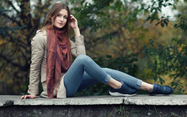 Brunette woman wearing a rust-colored scarf and casual outfit, sitting on a concrete ledge with a forest background, captured in a 4K Ultra HD desktop wallpaper style.