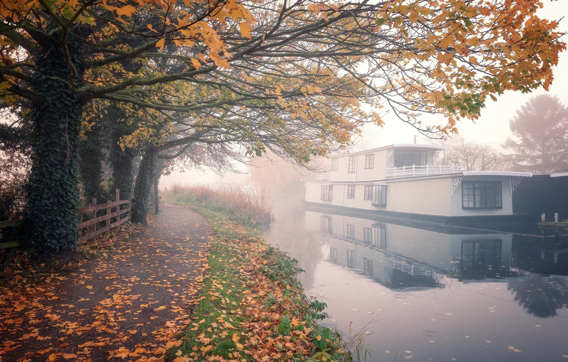 HD desktop wallpaper showing a tranquil fall scene with a canal reflecting a moored houseboat, surrounded by autumn leaves and misty atmosphere.