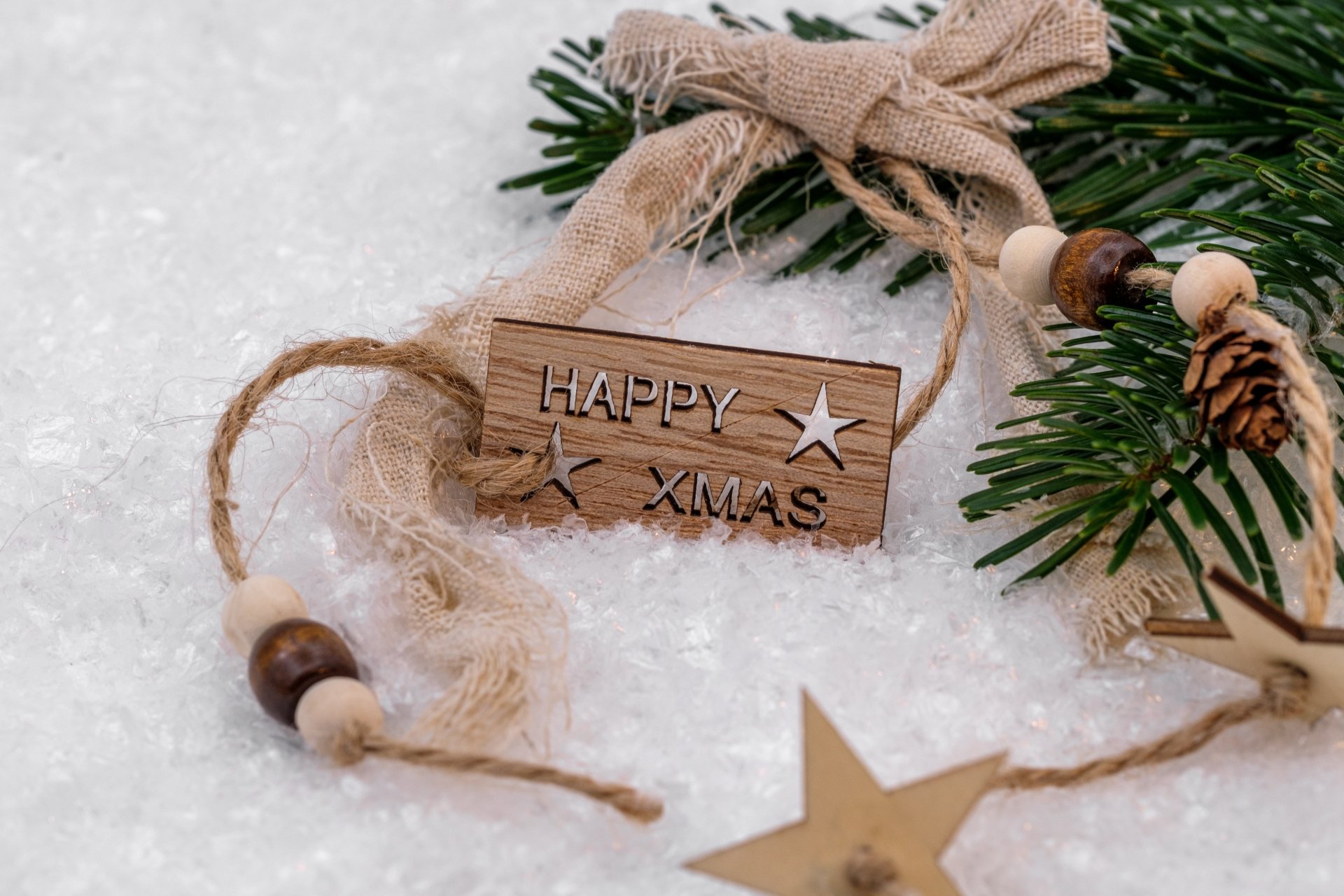5K Ultra HD PC desktop wallpaper of Christmas ornaments: a rustic 'HAPPY XMAS' wooden tag, star and bead decorations with pine sprig resting on snow, festive holiday scene.