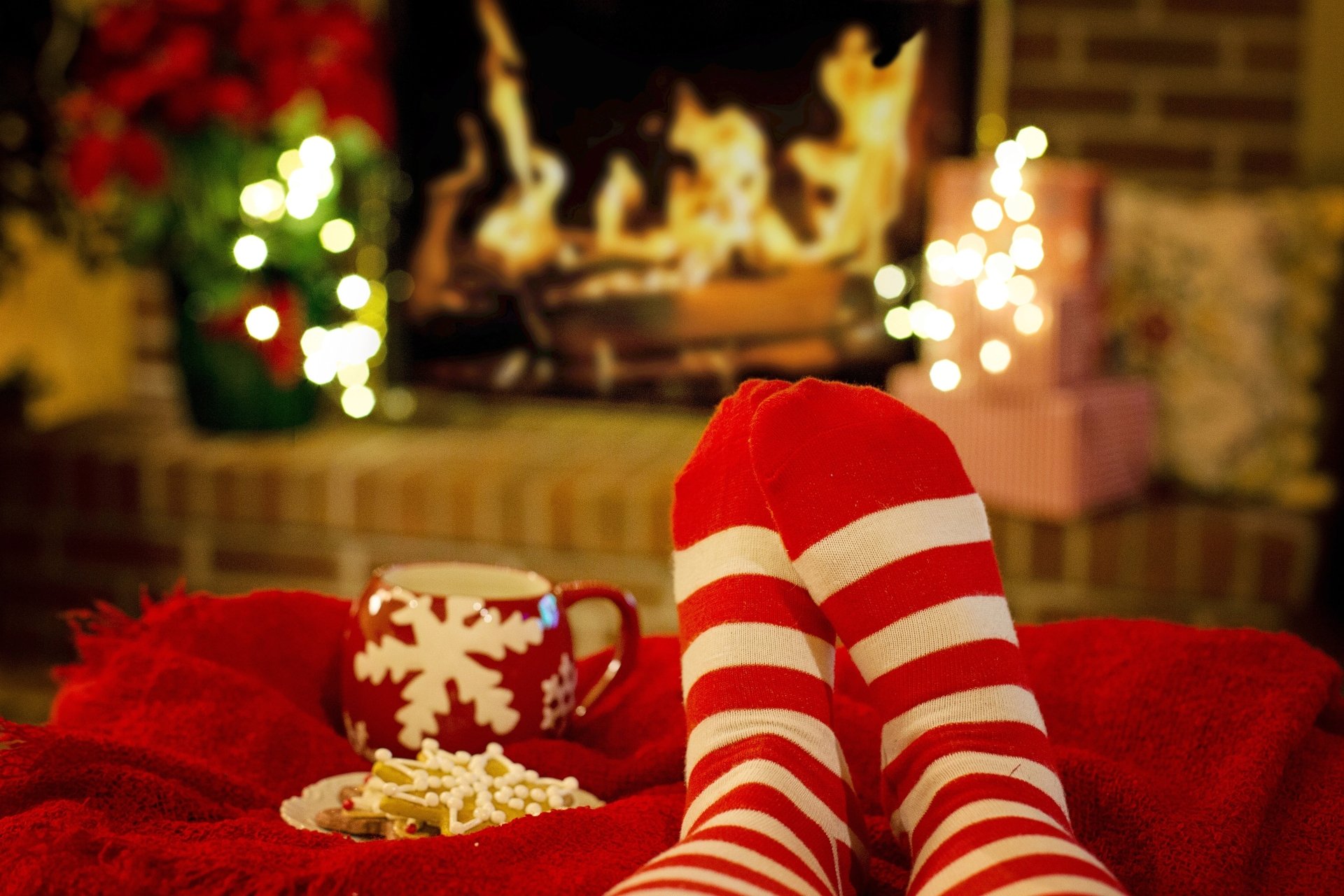 Cozy legs in red and white striped socks rest by a festive fireplace with a Christmas-themed mug and cookies, captured in vibrant 4K Ultra HD photography.