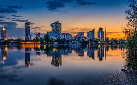 4K Ultra HD PC desktop wallpaper: Vienna skyline at sunset over a man-made riverfront in Austria, modern high-rises mirrored on calm water.