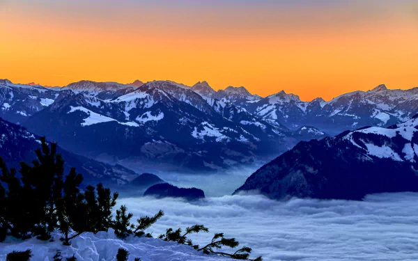 Snow-covered Bernese Alps in Switzerland at sunset with vibrant orange sky above mountainous peaks and a blanket of clouds below, captured in HD.