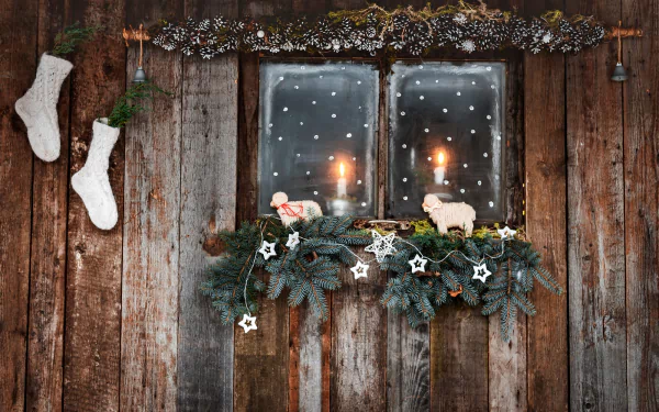 Cozy Christmas HD desktop wallpaper featuring a rustic wooden wall with stockings, ornaments, a candlelit window, and festive holiday greenery decorated with stars.