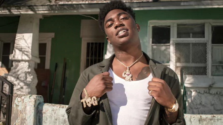 HD desktop wallpaper featuring a man in front of a house, wearing a necklace and adjusting his jacket.