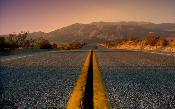 A scenic view of a road with a yellow center line stretches toward distant black mountains under a warm sky, framed by low bushes and the rugged terrain, creating a striking wallpaper backdrop.